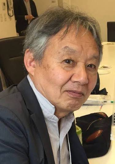 Satoru Okada, grey-haired, in a dark jacket over a light shirt, seated at a table and looking towards the camera.
