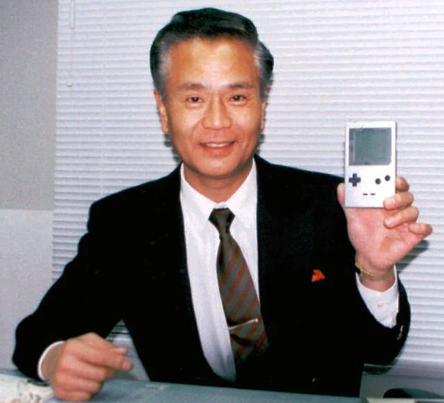 Gunpei Yokoi seated at a desk in a dark suit, smiling and holding up a silver Game Boy Pocket.