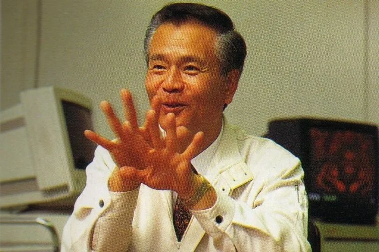 Gunpei Yokoi in a white lab coat, smiling and gesturing with both hands raised, in an office with computer monitors behind him.